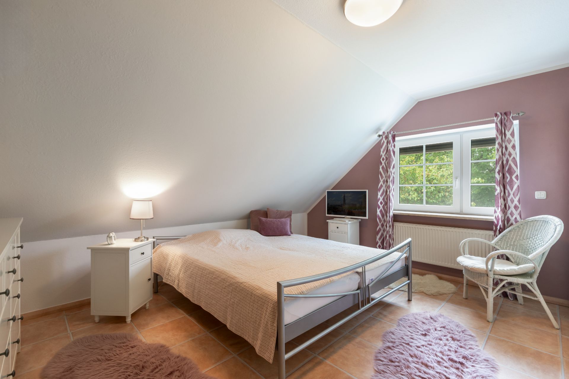 Helles Schlafzimmer mit Doppelbett, Nachttischen, Sessel und Fenster mit rosa Vorhängen.