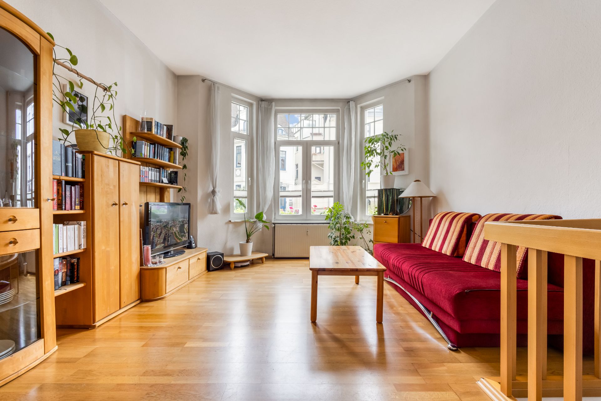 Helles Wohnzimmer mit rotem Sofa, Holzmöbeln, Pflanzen und großem Erkerfenster.