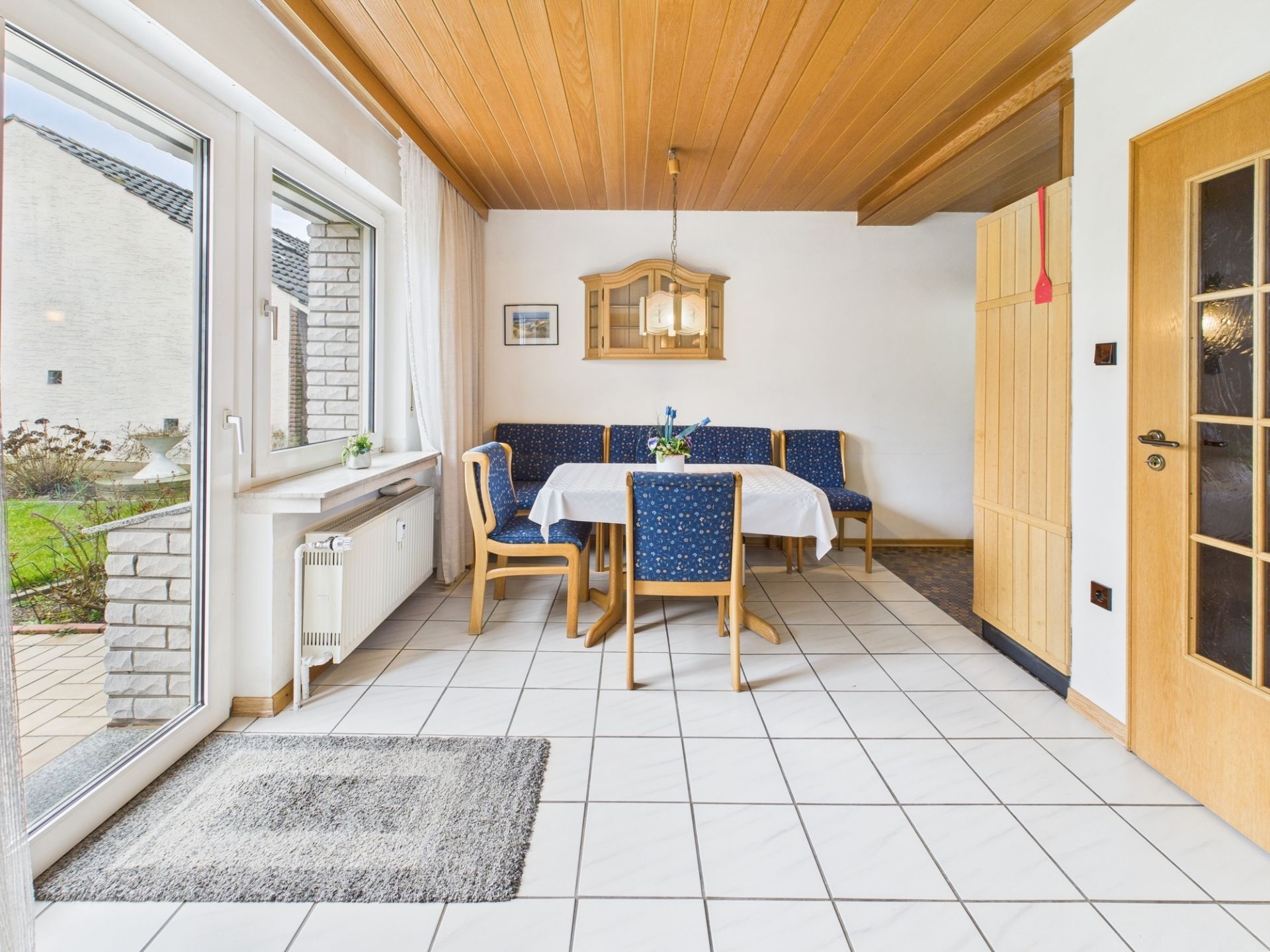 Essbereich mit Eckbank und Holztisch vor einem großen Fenster, Fliesenboden und Holdecken.