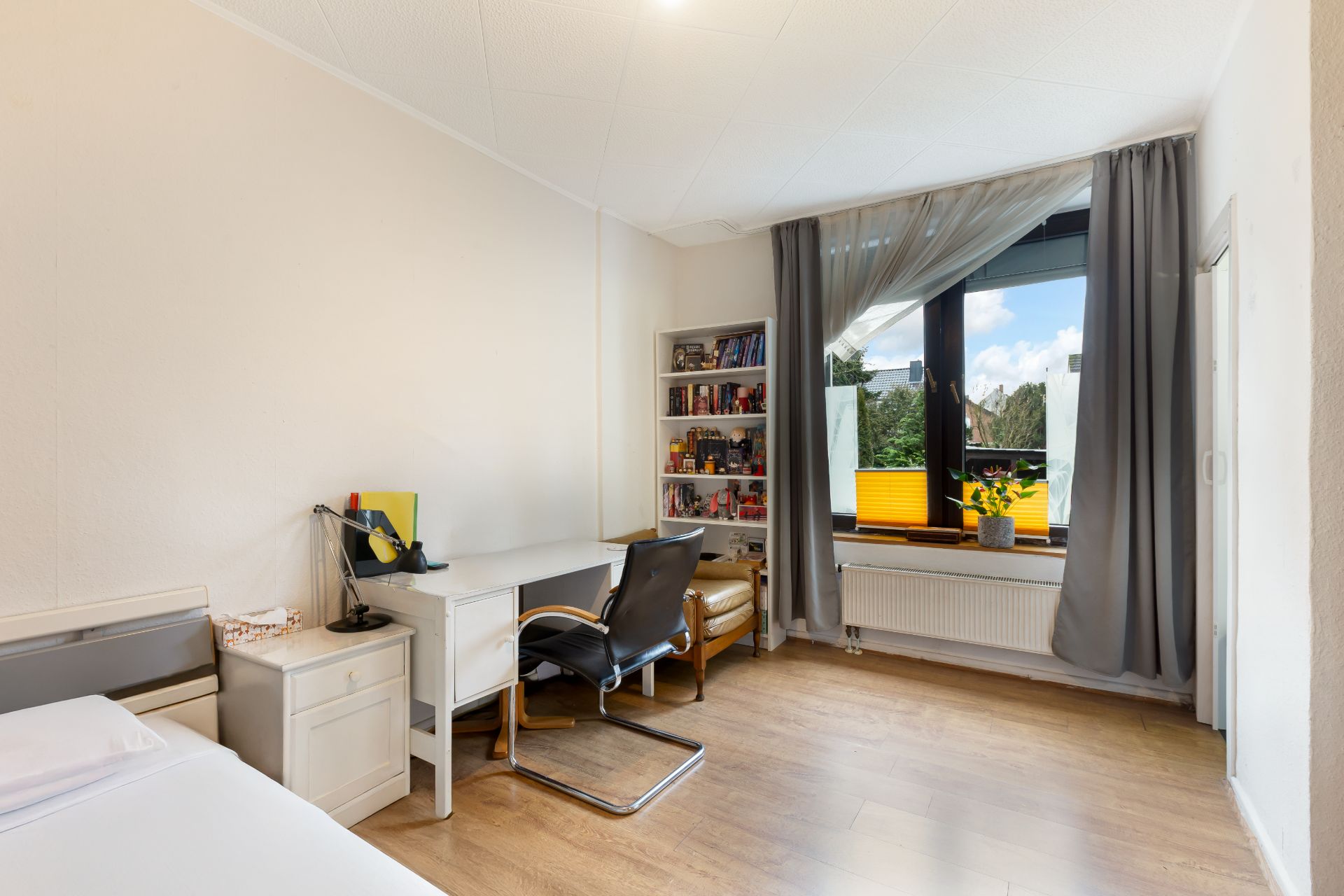 Helles Zimmer mit Einzelbett, Schreibtisch, Bücherregal und großem Fenster mit Vorhängen.