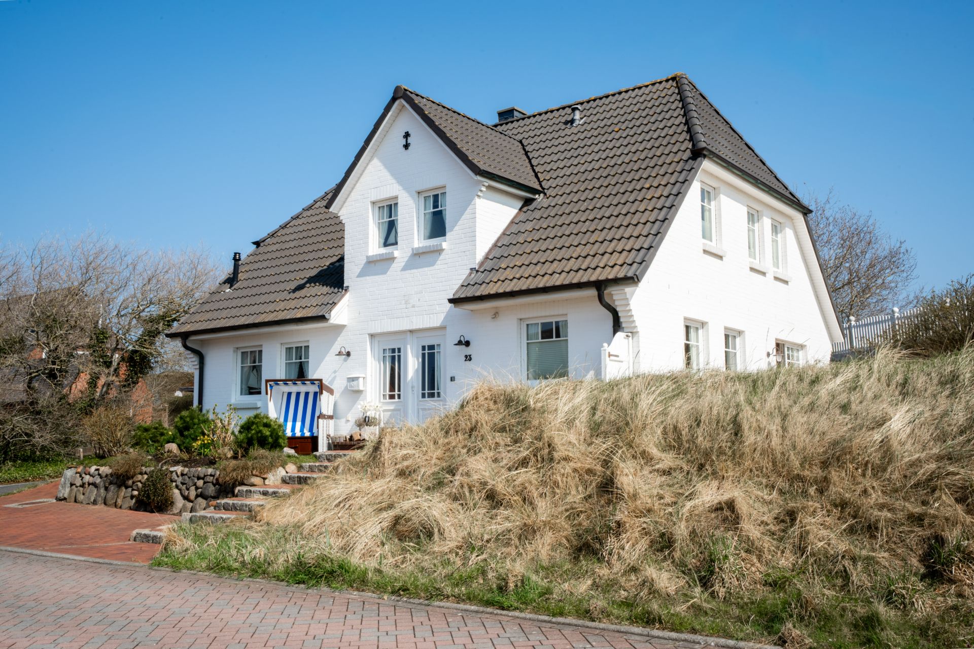 Weißes Einfamilienhaus mit dunklem Dach und kleinem Garten vorne, blauer Himmel.