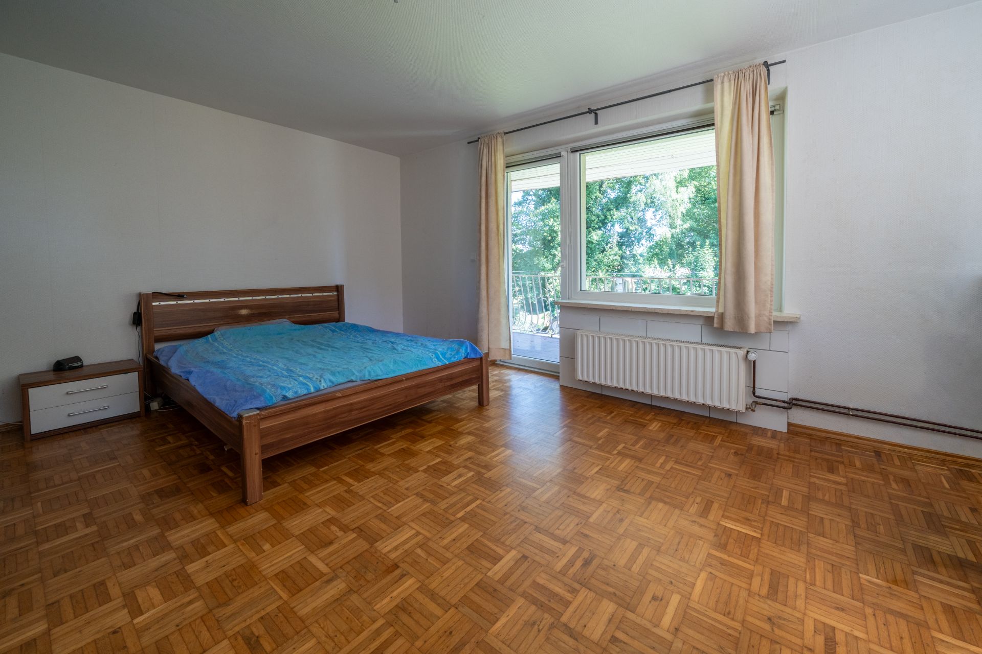 Geräumiges Schlafzimmer mit Holzboden, Doppelbett, Nachttisch und großen Fenstern mit Blick ins Grüne.