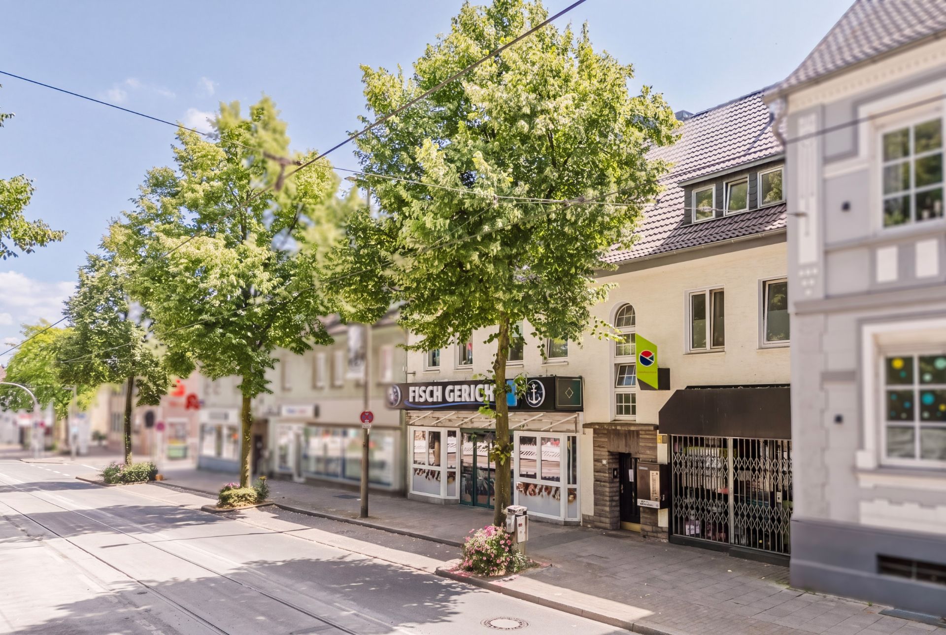 Straßenszene mit Bäumen und Geschäften, darunter ein Fischrestaurant, und Wohnhäusern im Hintergrund.