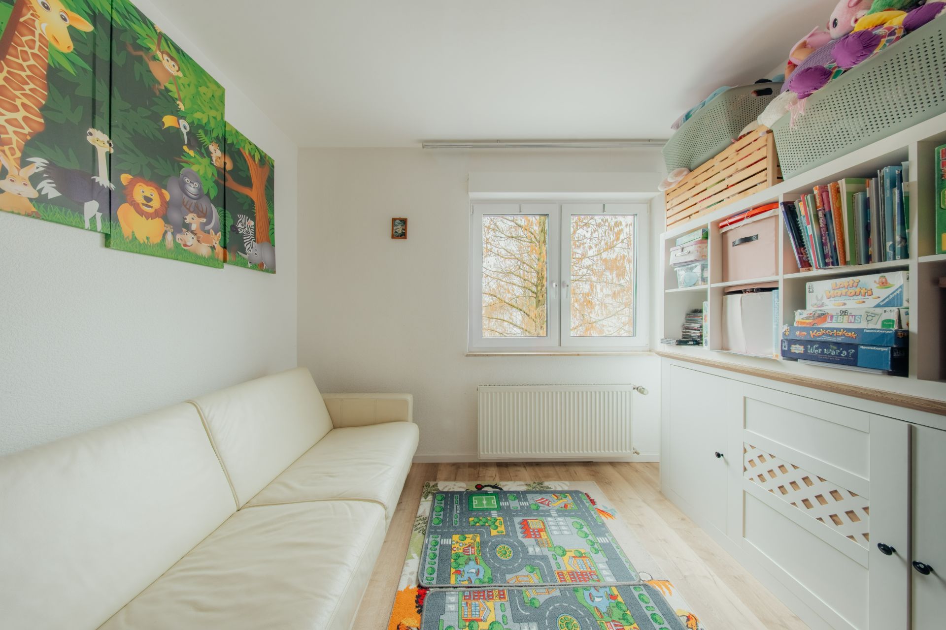 Kinderzimmer mit weißem Sofa, buntem Spielteppich, Bücherregal und Fenster.
