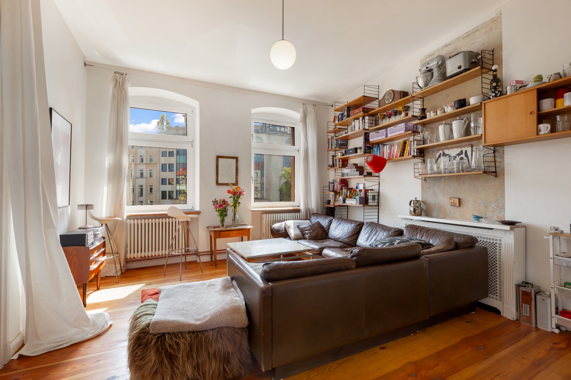 Helles Wohnzimmer mit branem Sofa, Bücherregalen und zwei großen Fenstern.