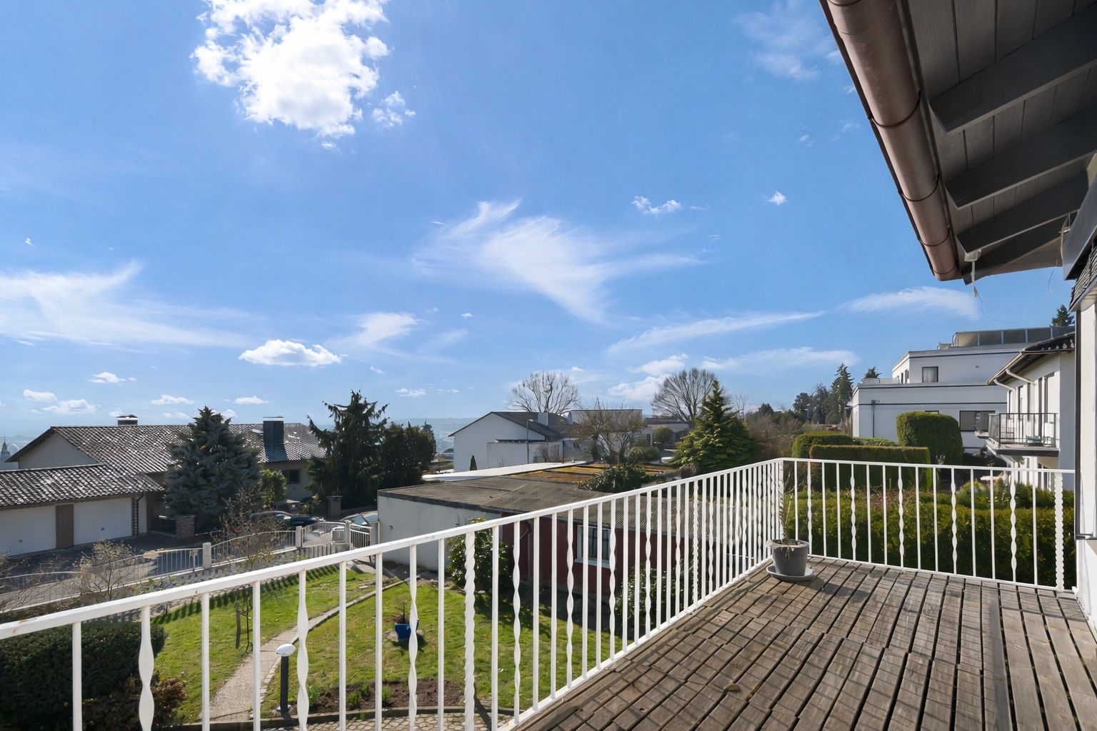 Balkon mit Holzboden und weißem Geländer, Blick auf Nachbarhäuser und Garten.