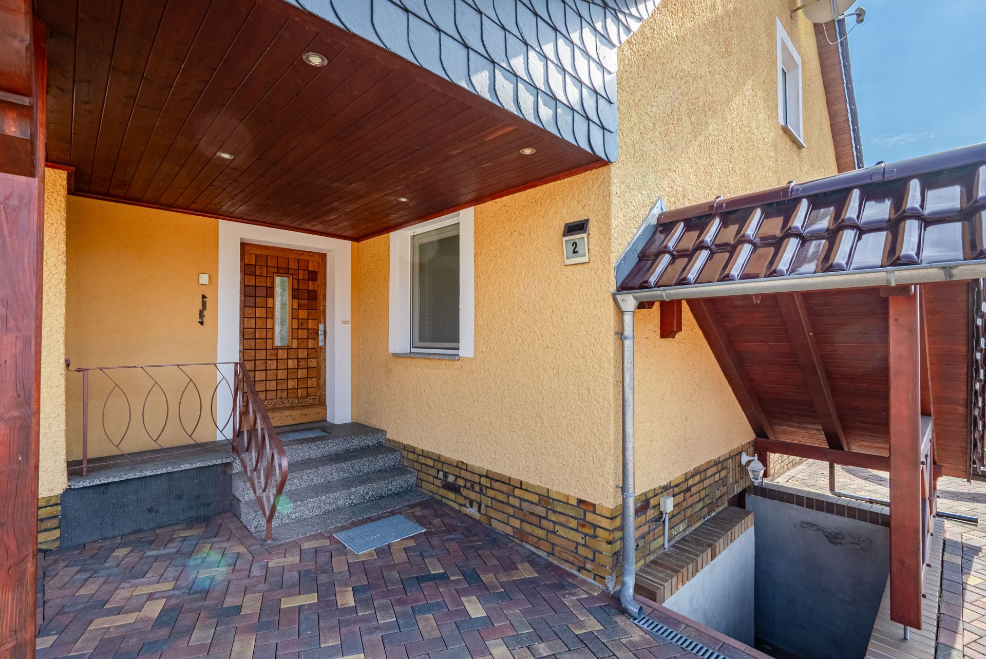 Eingangsbereich eines Hauses mit Treppe, Tür, Fenster und überdachtem Kellerabgang.