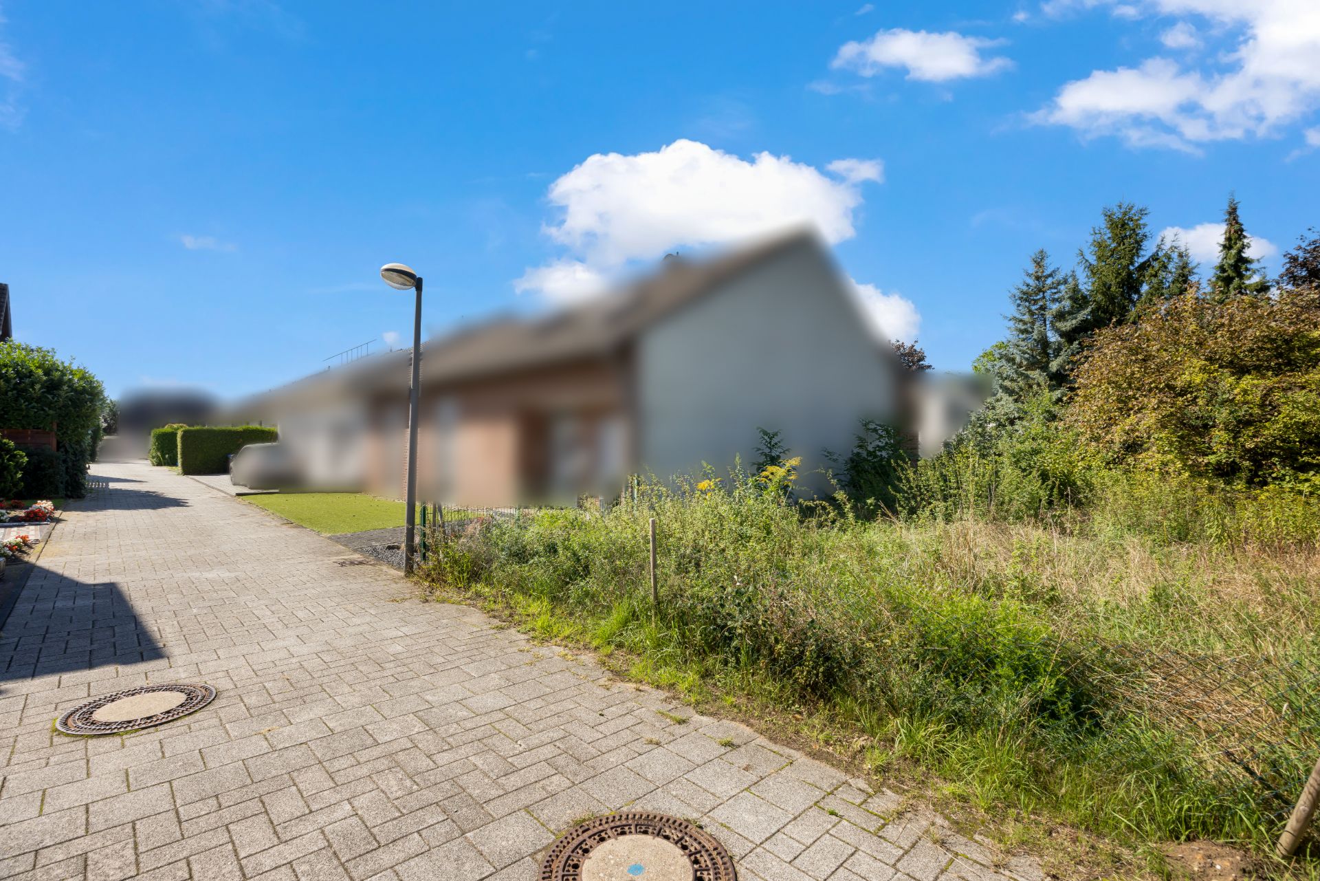 Gepflasterter Gehweg neben einer Wiese mit Büschen, im Hintergrund ein Wohnhaus.