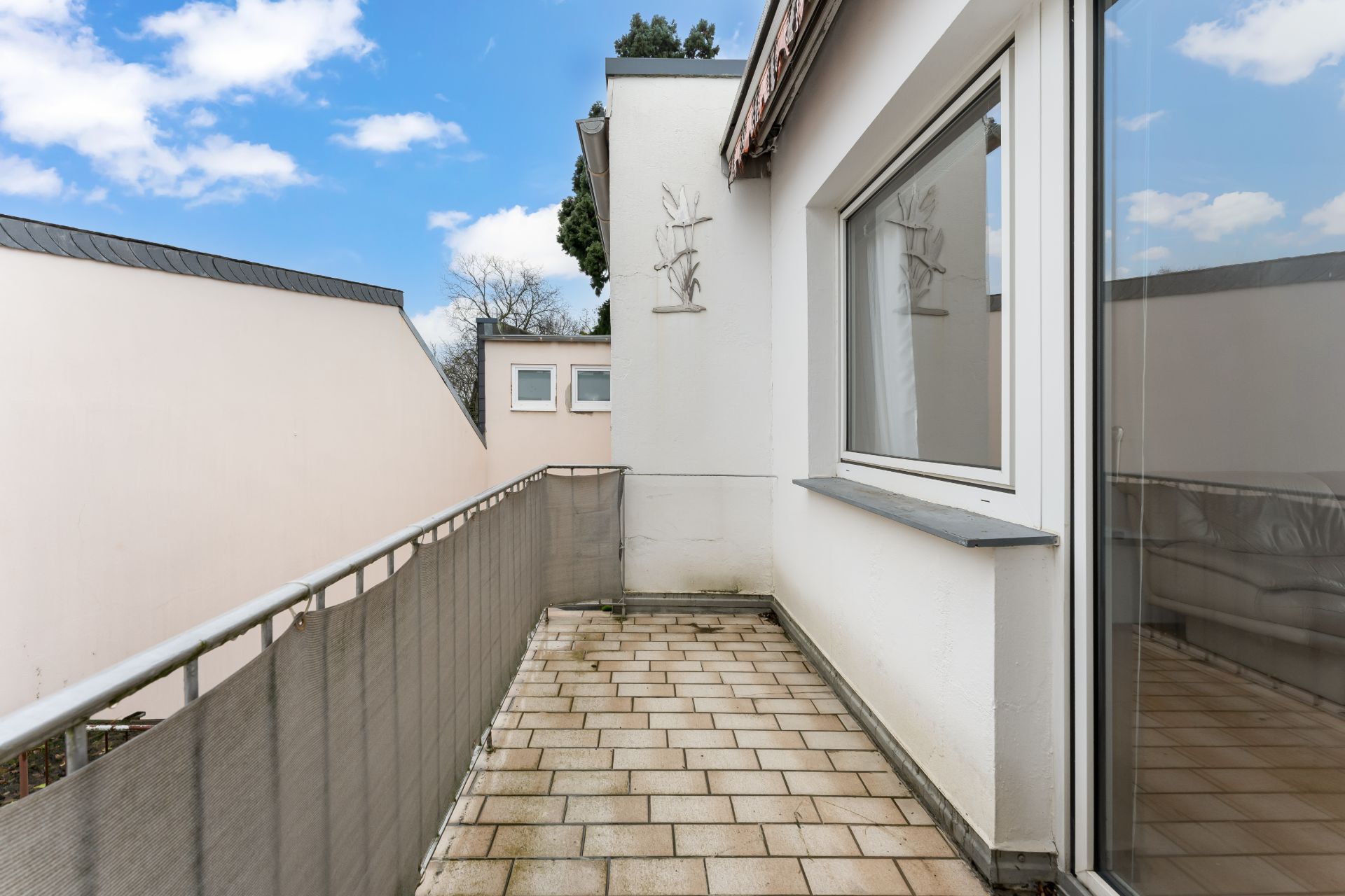 Gefliester Balkon mit Metallgeländer und Blick auf Nachbargebäude, Fenster und Glastür sichtbar.