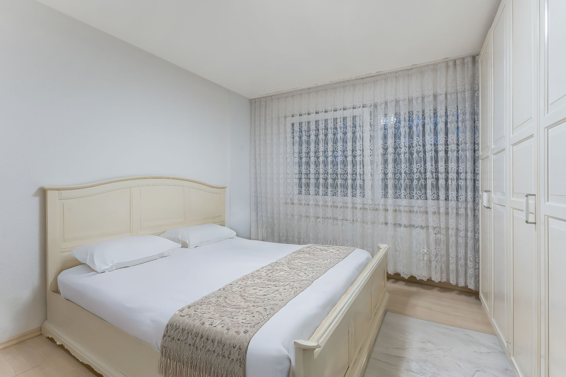 Helles Schlafzimmer mit Doppelbett, weißem Kleiderschrank und großen Fenstern mit Gardinen.