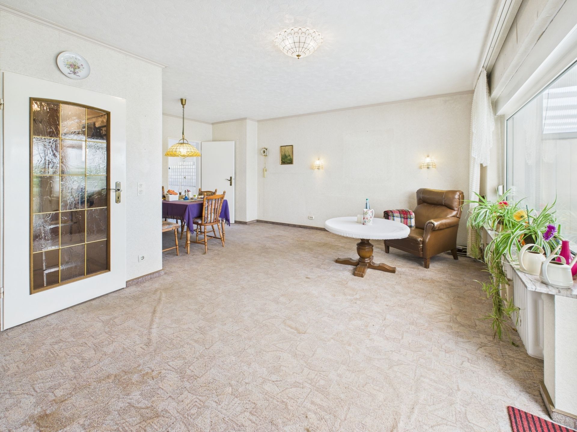 Helles Wohnzimmer mit Sessel, Tisch, Essbereich und vielen Pflanzen am Fenster.