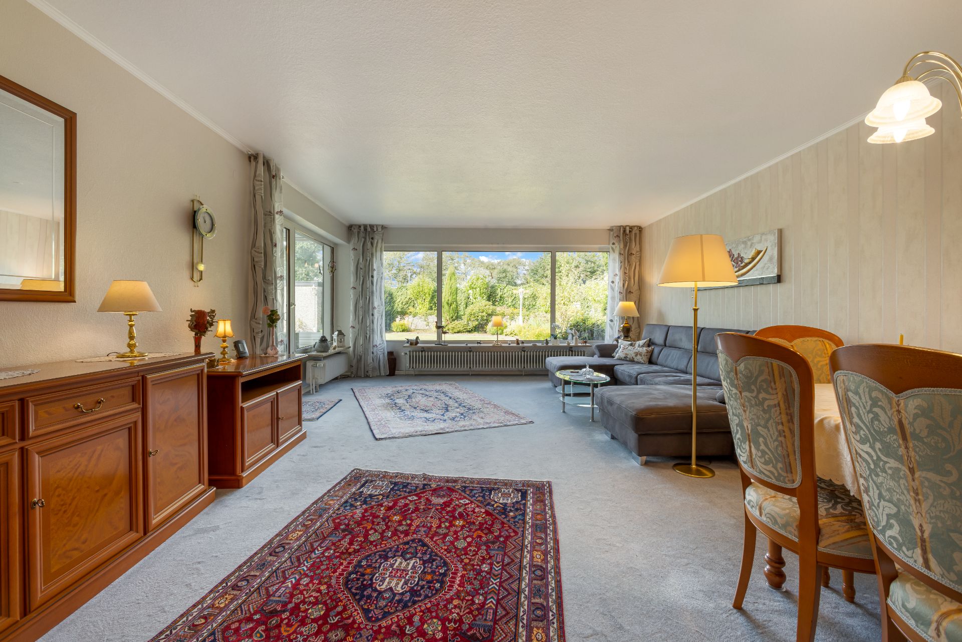 Helles Wohnzimmer mit großen Fenstern, Teppichen, Sofa, Esstisch und Holzmöbeln.