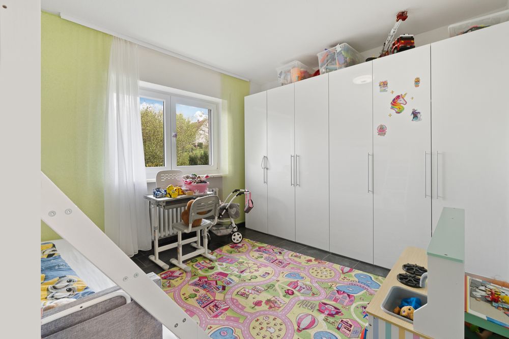Helles Kinderzimmer mit buntem Teppich, Schreibtisch am Fenster und weißen Kleiderschränken.