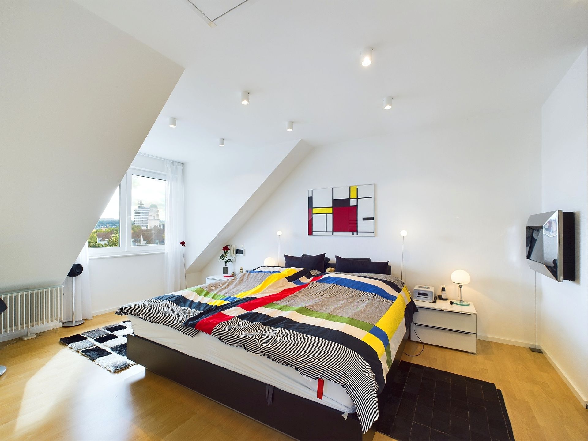 Modernes Schlafzimmer mit buntem Bettbezug, weißen Wänden und großem Fenster.