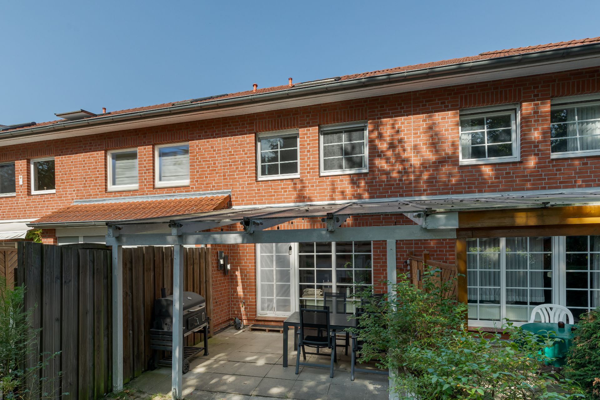 Backstein-Reihenhaus mit Terrasse, Gartenmöbeln und Grill unter einer Pergola.