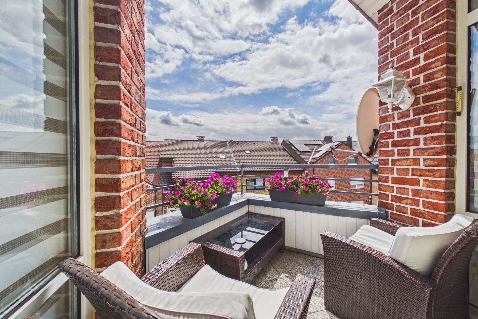 Gemütlicher Balkon mit zwei Sesseln, Tisch, Blumentöpfen und Blick auf Nachbarhäuser.
