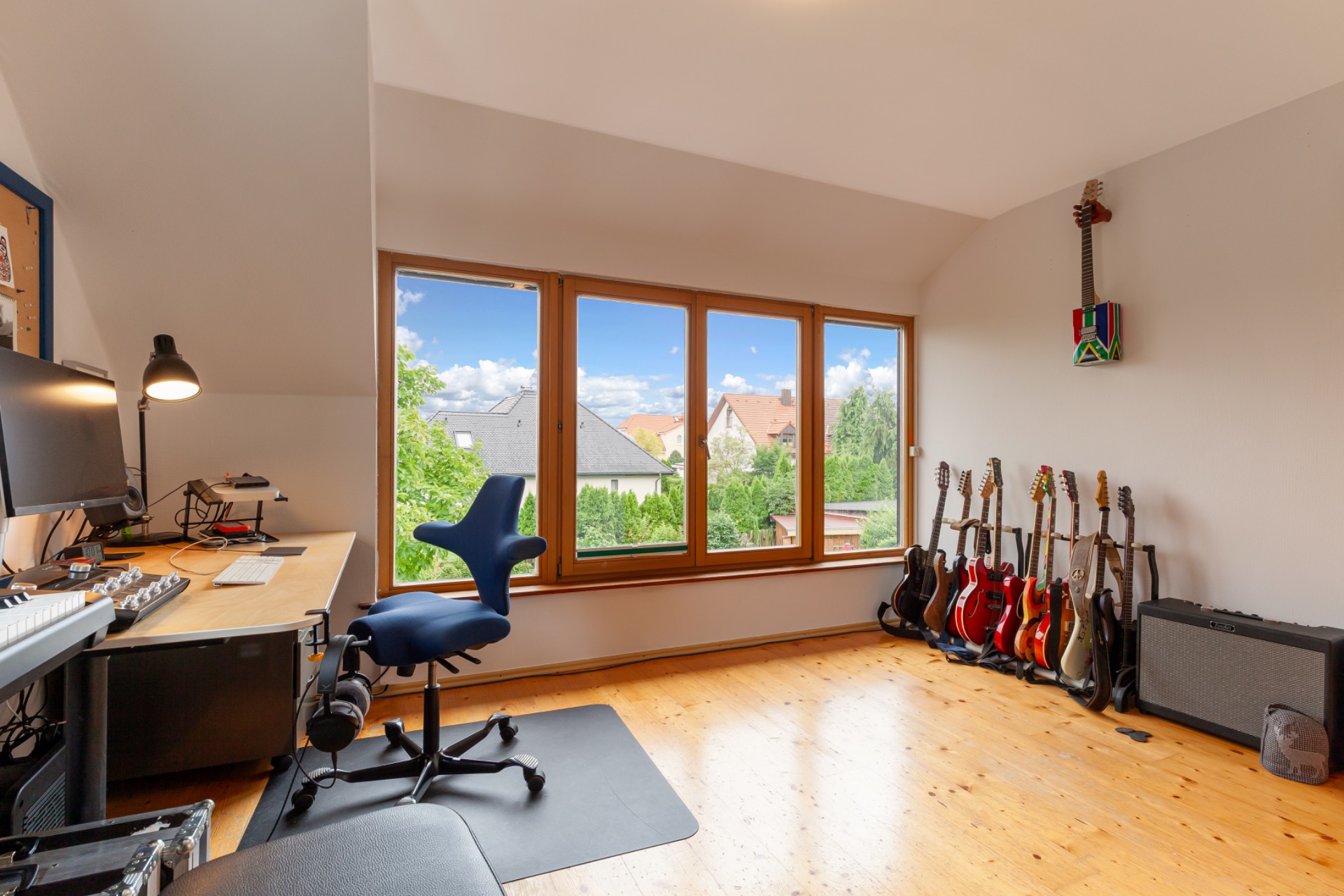 Helles Zimmer mit großem Fenster, Schreibtisch, Bürostuhl und mehreren Gitarren an der Wand.