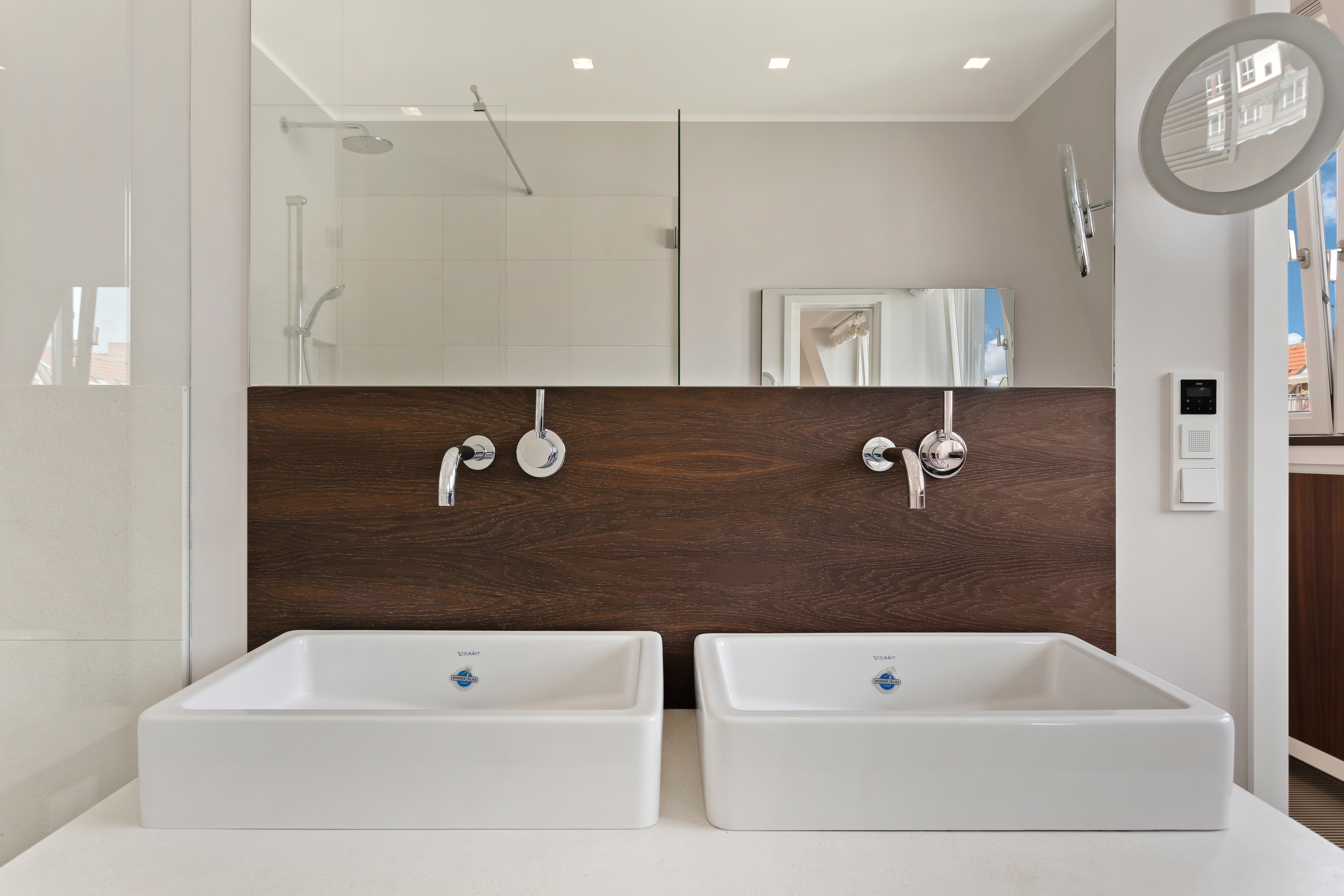 Zwei moderne weiße Waschbecken mit Wandarmaturen vor Holzverkleidung im Badezimmer.
