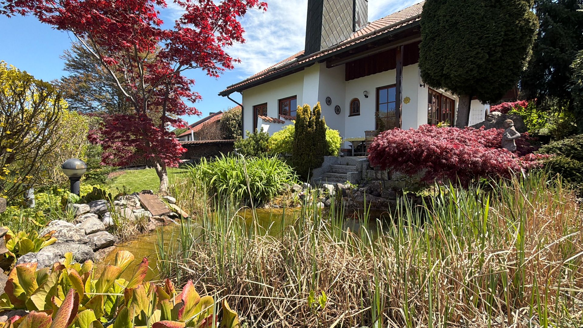 Weißes Wohnhaus mit Garten, Teich, Steinweg und bunten Büschen im Vordergrund.