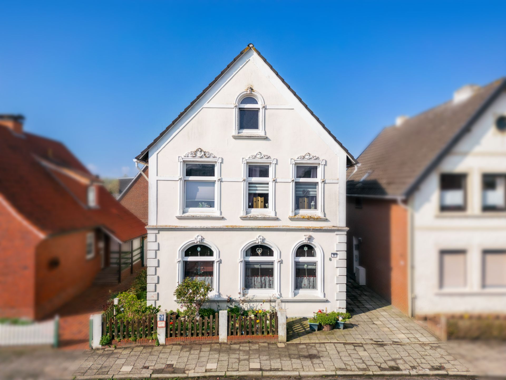 Zweistöckiges weißes Wohnhaus mit dekorativen Fenstern und kleinem Garten vorne.