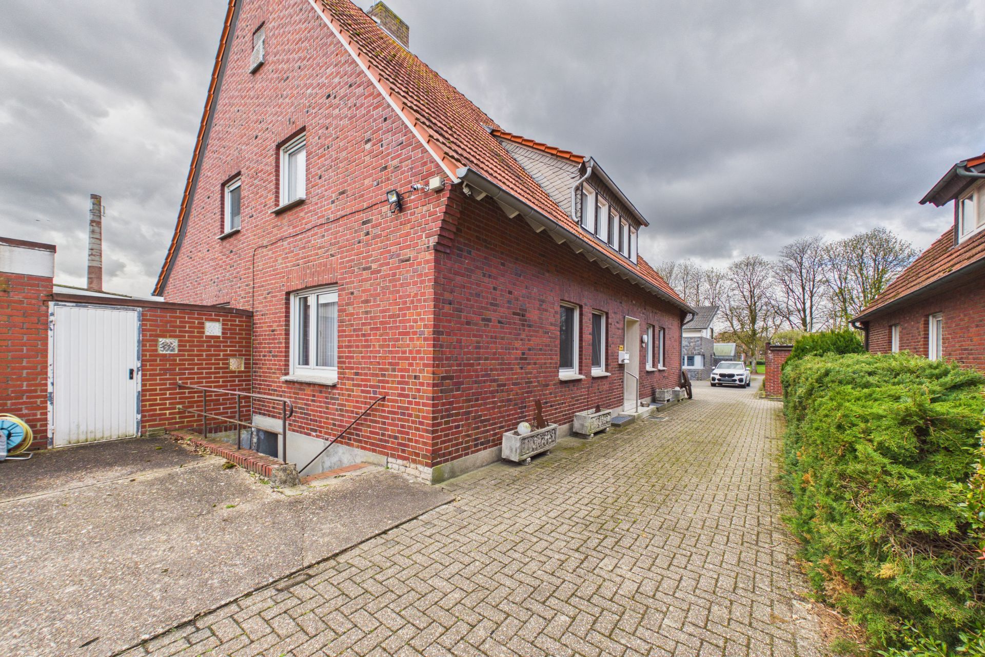 Backsteinwohnhaus mit rotem Dach und gepflasterter Einfahrt, Büsche rechts.