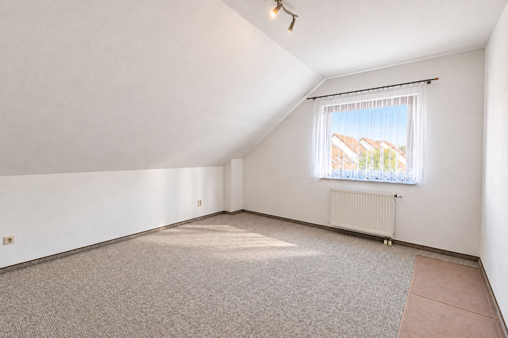 Helles Dachzimmer mit Teppichboden, Fenster mit Vorhang und Heizkörper unter dem Fenster.