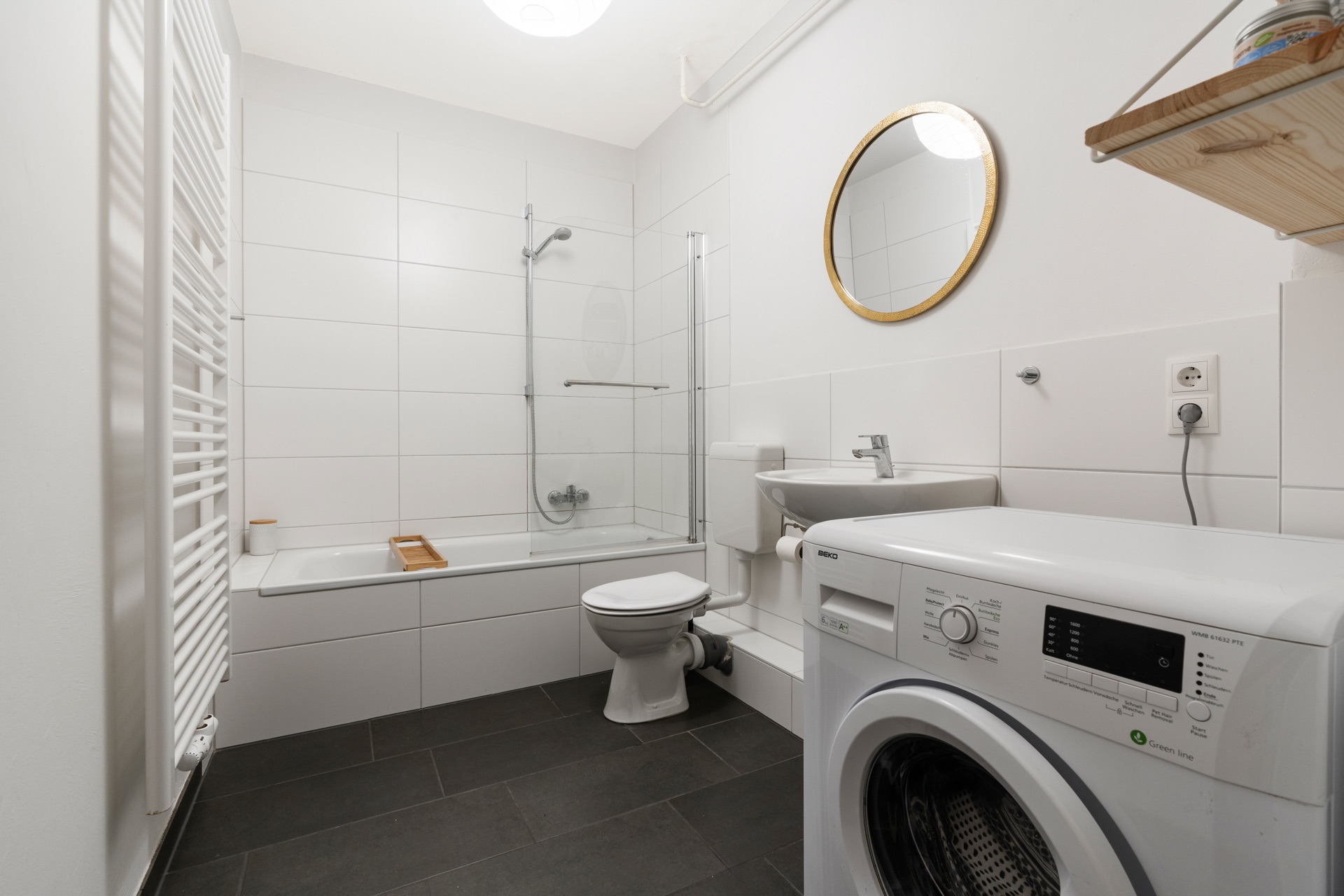 Helles Bad mit Badewanne, Duschwand, WC, Waschbecken, Waschmaschine und rundem Spiegel.