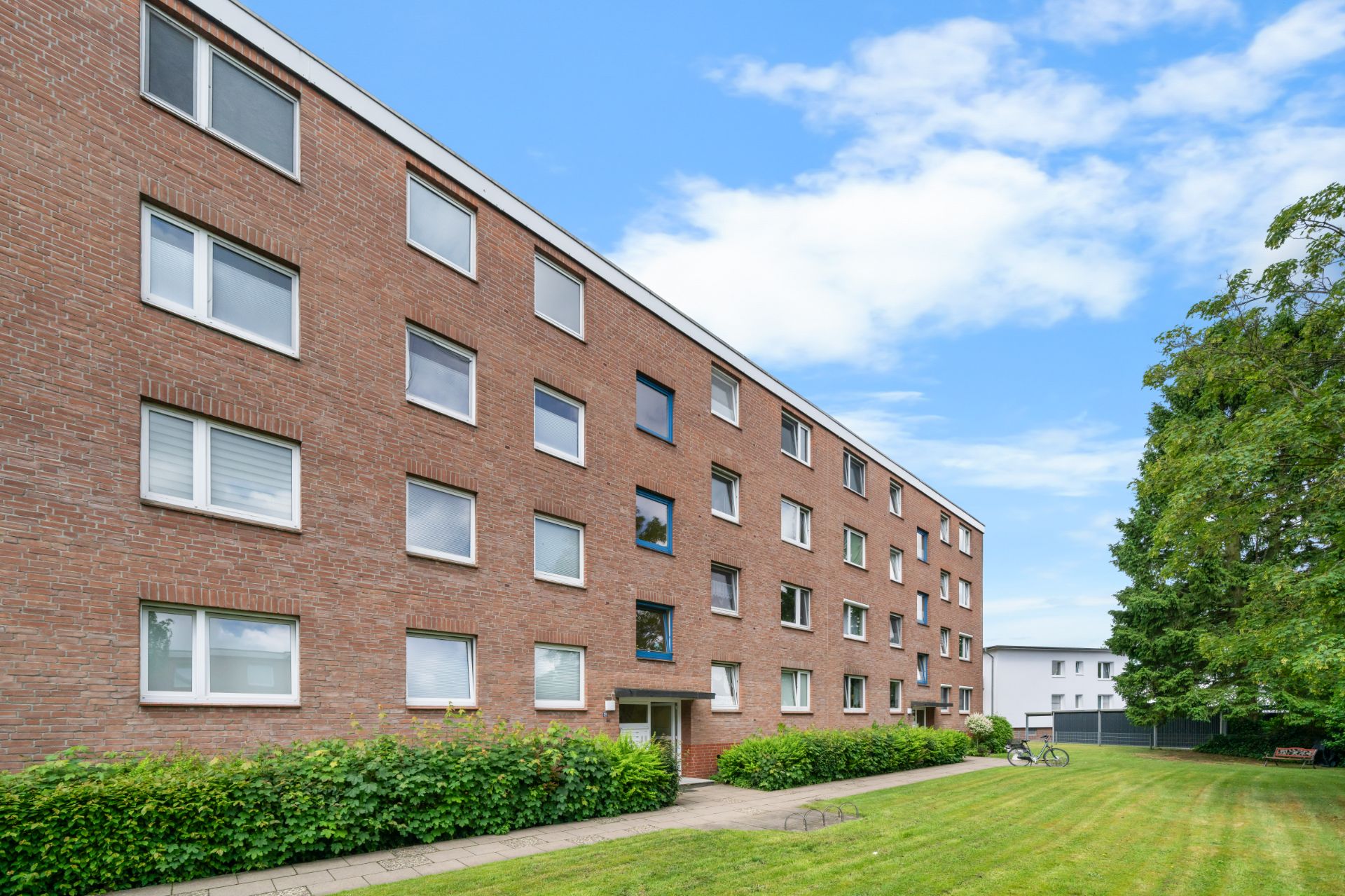 Mehrstöckiges Wohngebäude aus rotem Backstein mit vielen Fenstern und grünem Vorgarten.