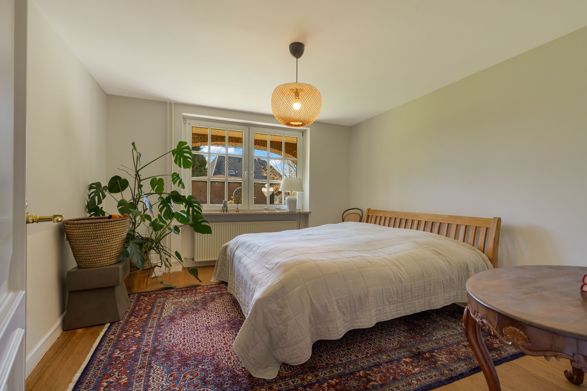 Schlafzimmer mit Doppelbett, großem Fenster, Zimmerpflanze und Teppich auf Holzboden.