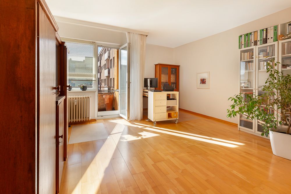 Helles Wohnzimmer mit Holzfußboden, großen Fenstern, Regal, Pflanzen und Zugang zum Balkon.