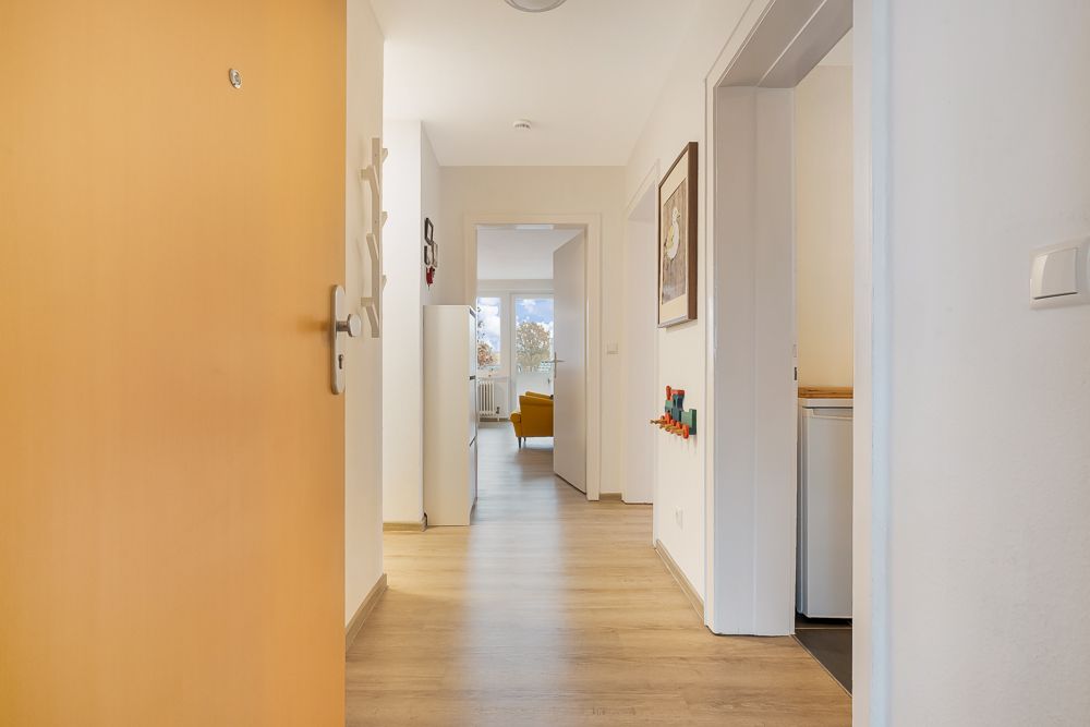 Heller Flur mit Holzboden, mehreren Türen und Blick auf ein gelbes Sofa im Wohnzimmer.