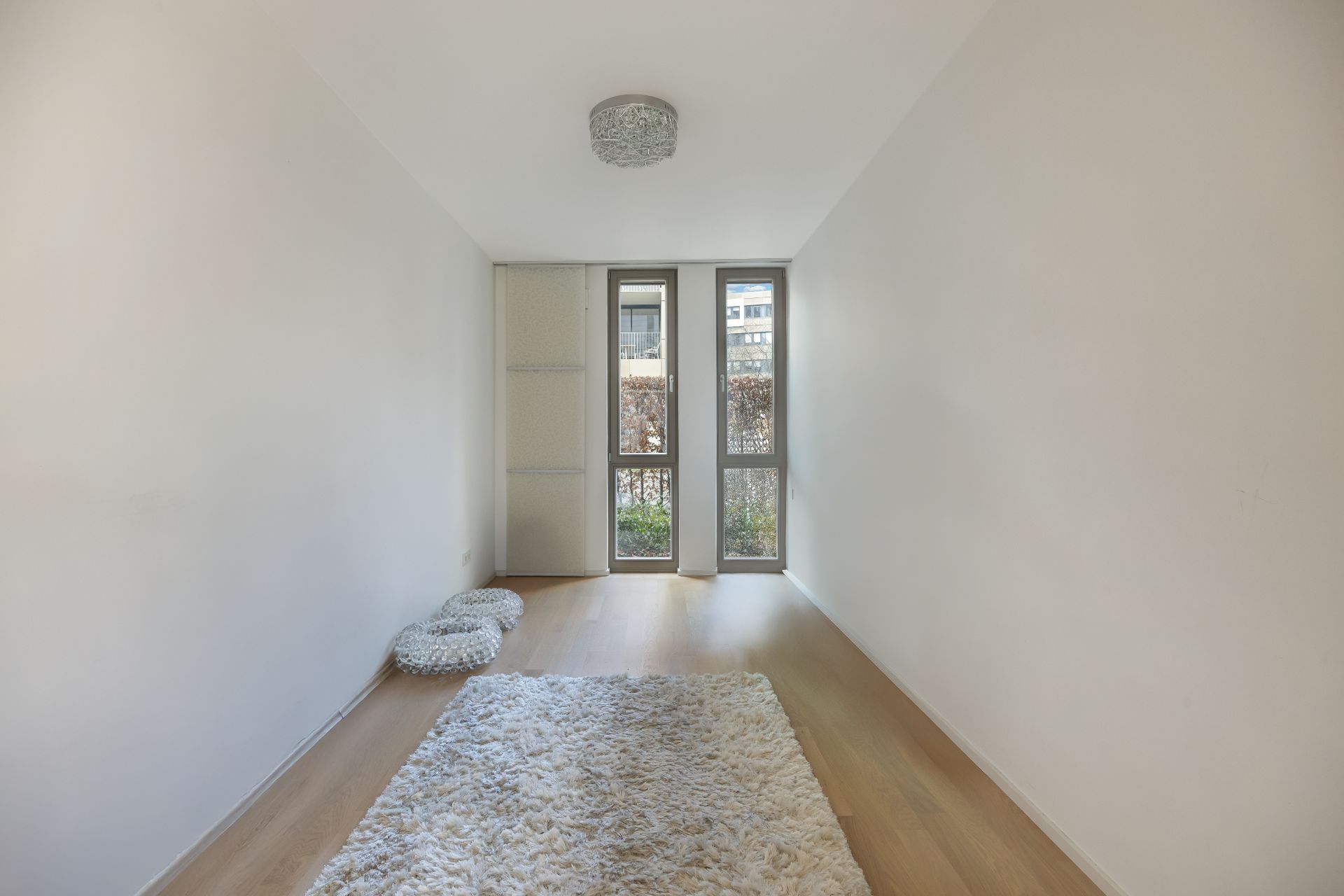 Heller, leerer Raum mit Holzboden, Teppich und großen Fenstern.