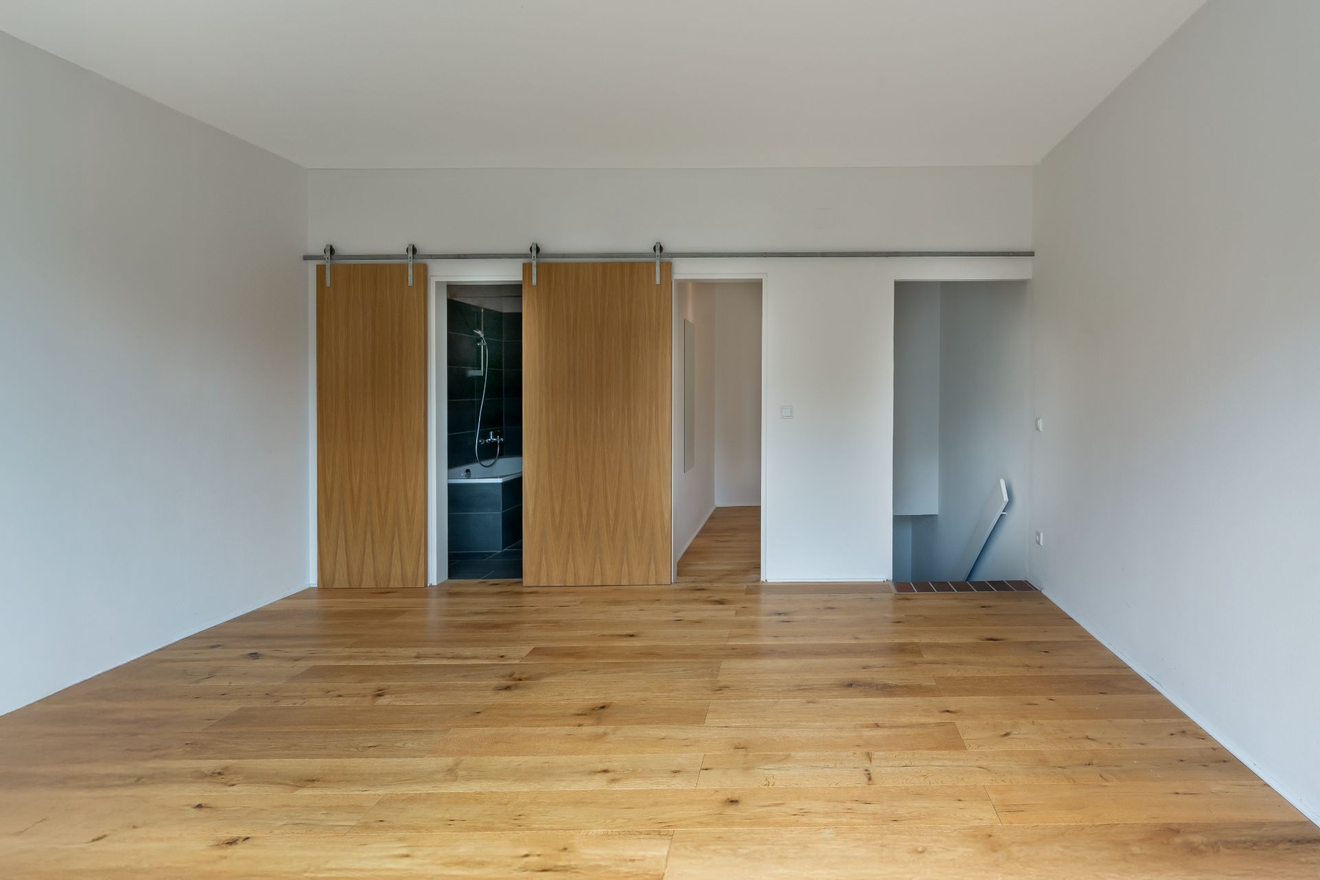 Leerer Raum mit Holzfußboden, zwei Schiebetüren, offenem Bad und Treppe nach unten.