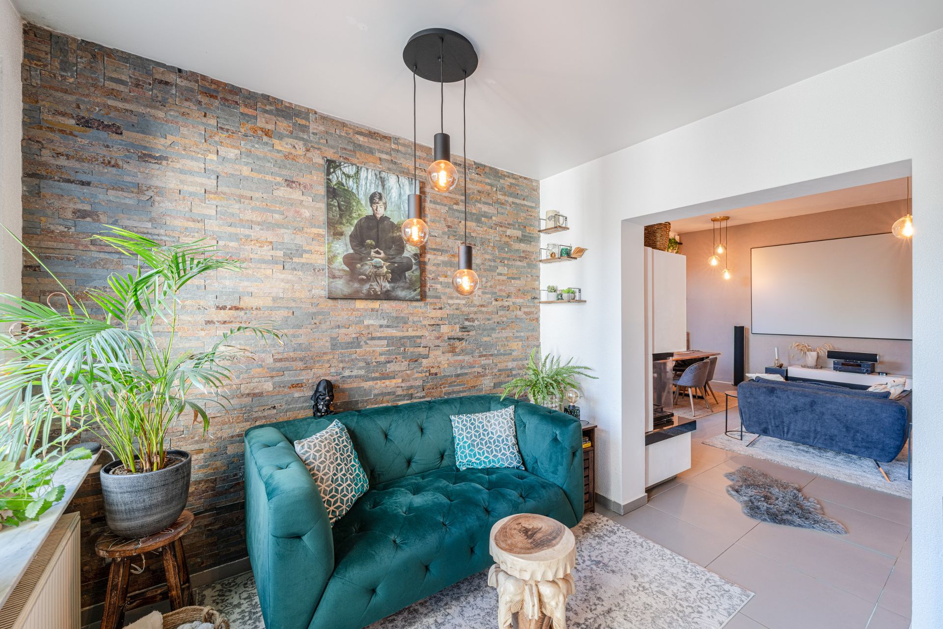 Helles Wohnzimmer mit grüner Couch, Pflanzen, gemauerter Wand und Blick in ein Ess- und Wohnzimmer.