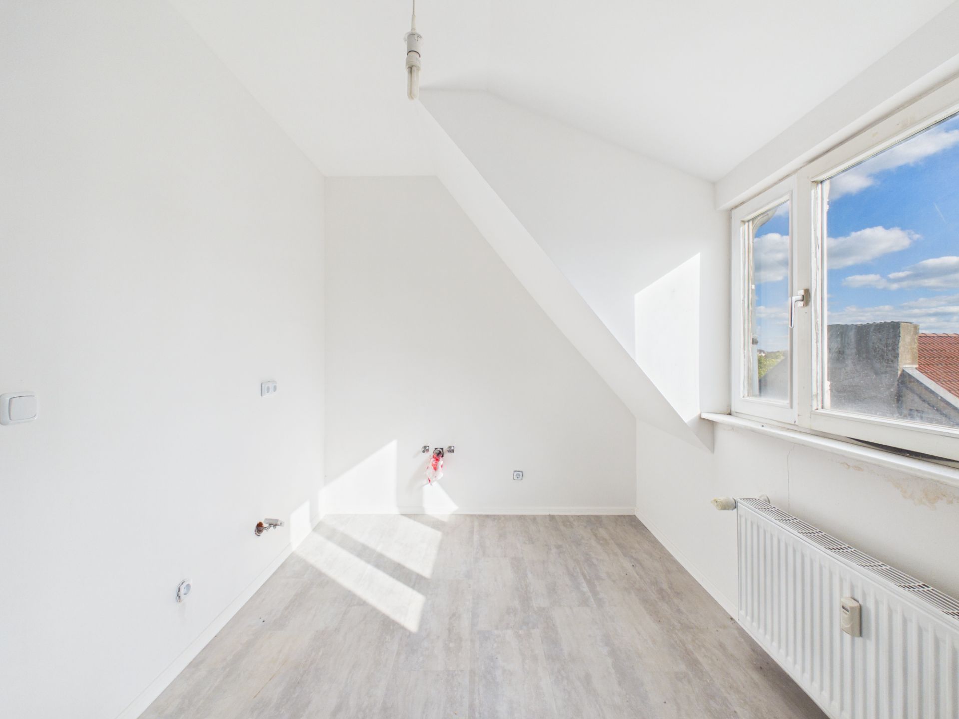 Heller, leerer Raum mit schräger Decke, großem Fenster und Heizkörper unter dem Fenster.