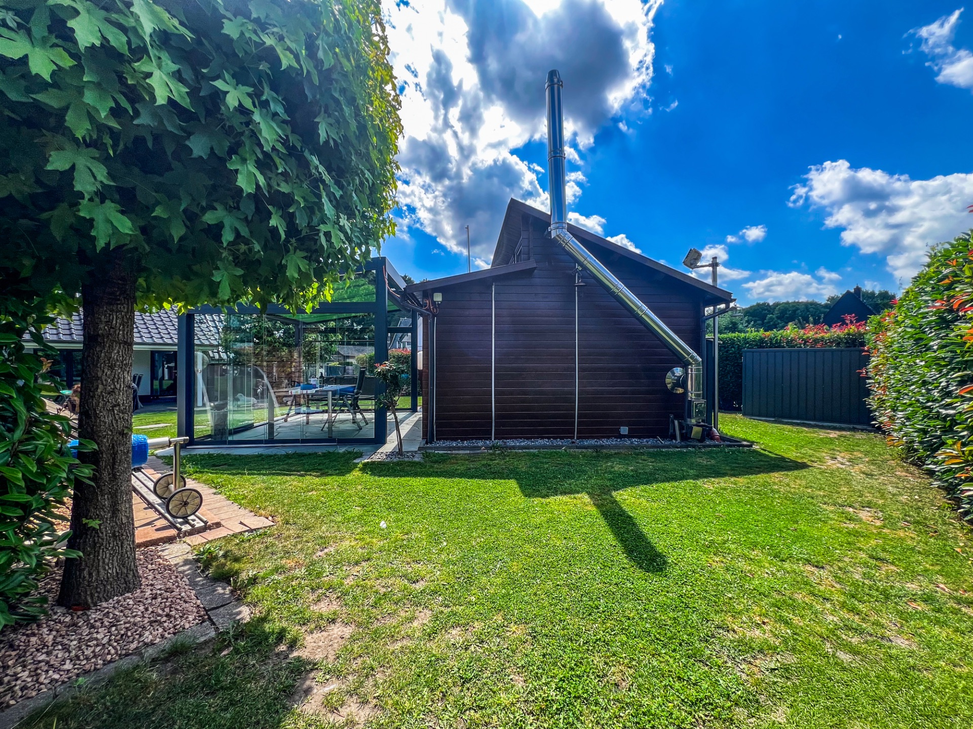 Garten mit Rasen, Baum, Holzhaus und überdachter Terrasse mit Sitzbereich.