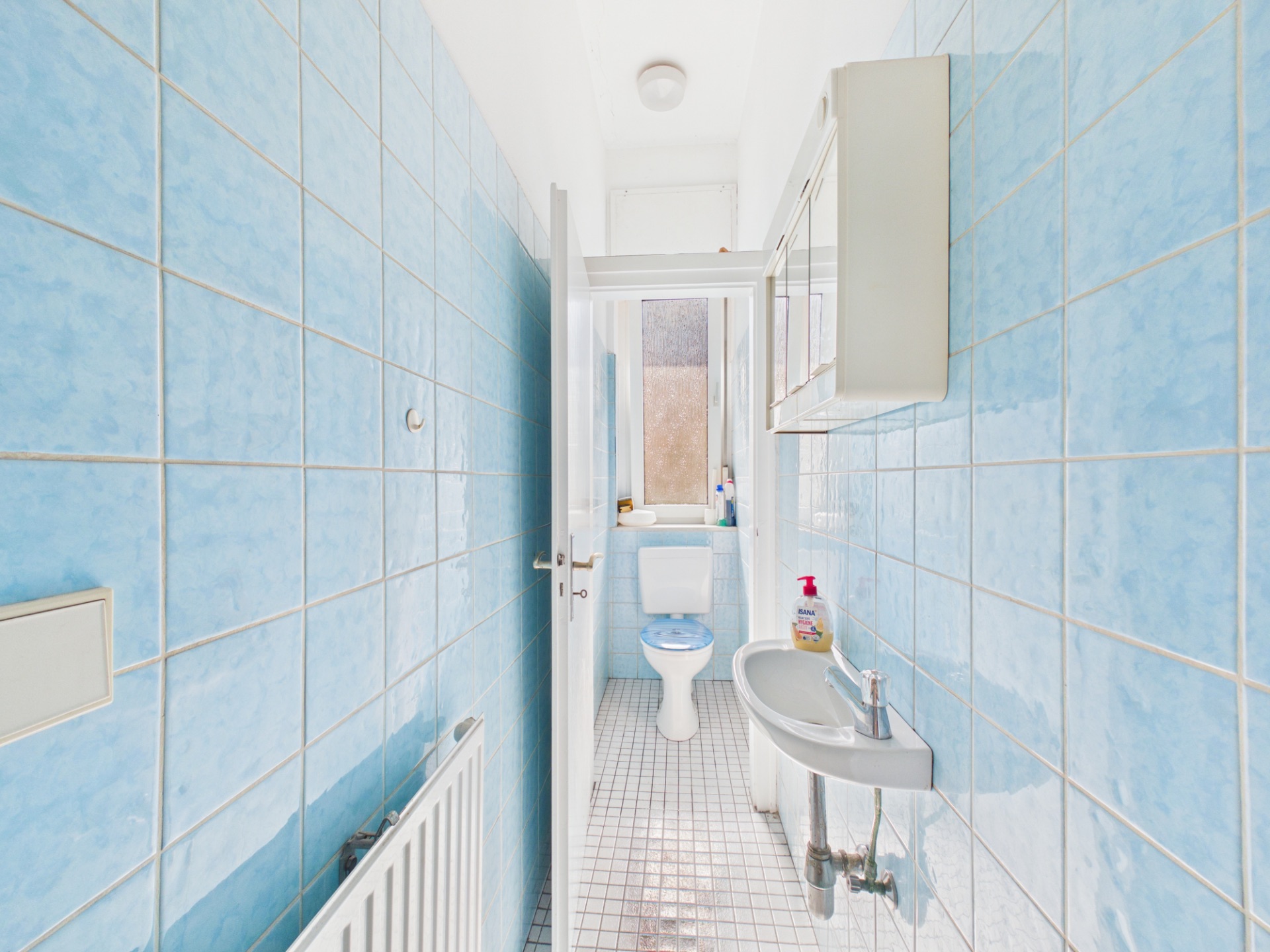 Schmales Badezimmer mit blauen Fliesen, WC, Waschbecken und Fenster am Ende.