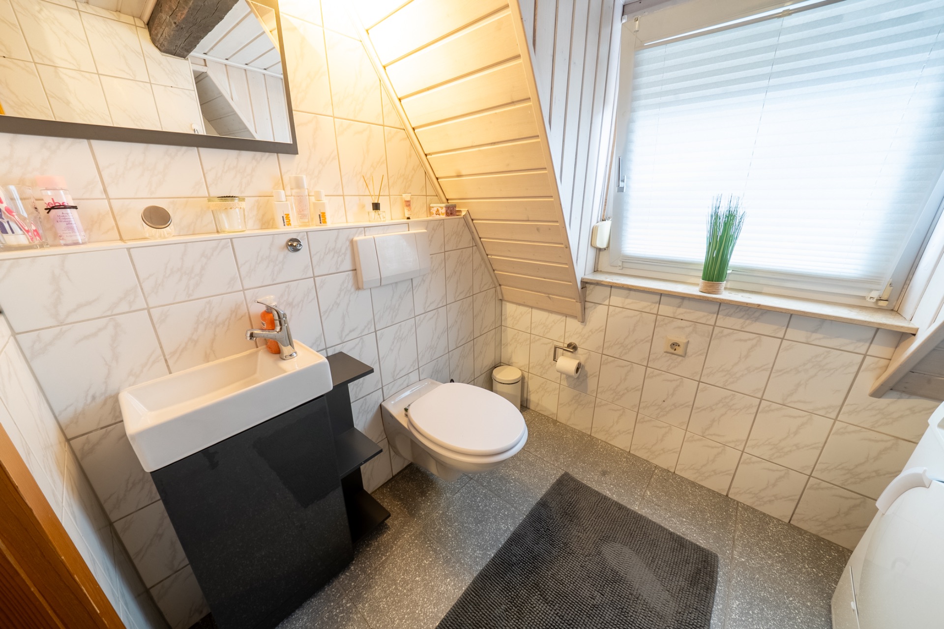 Kleines Badezimmer mit Waschbecken, Wandtoilette, Fenster und Badvorleger.