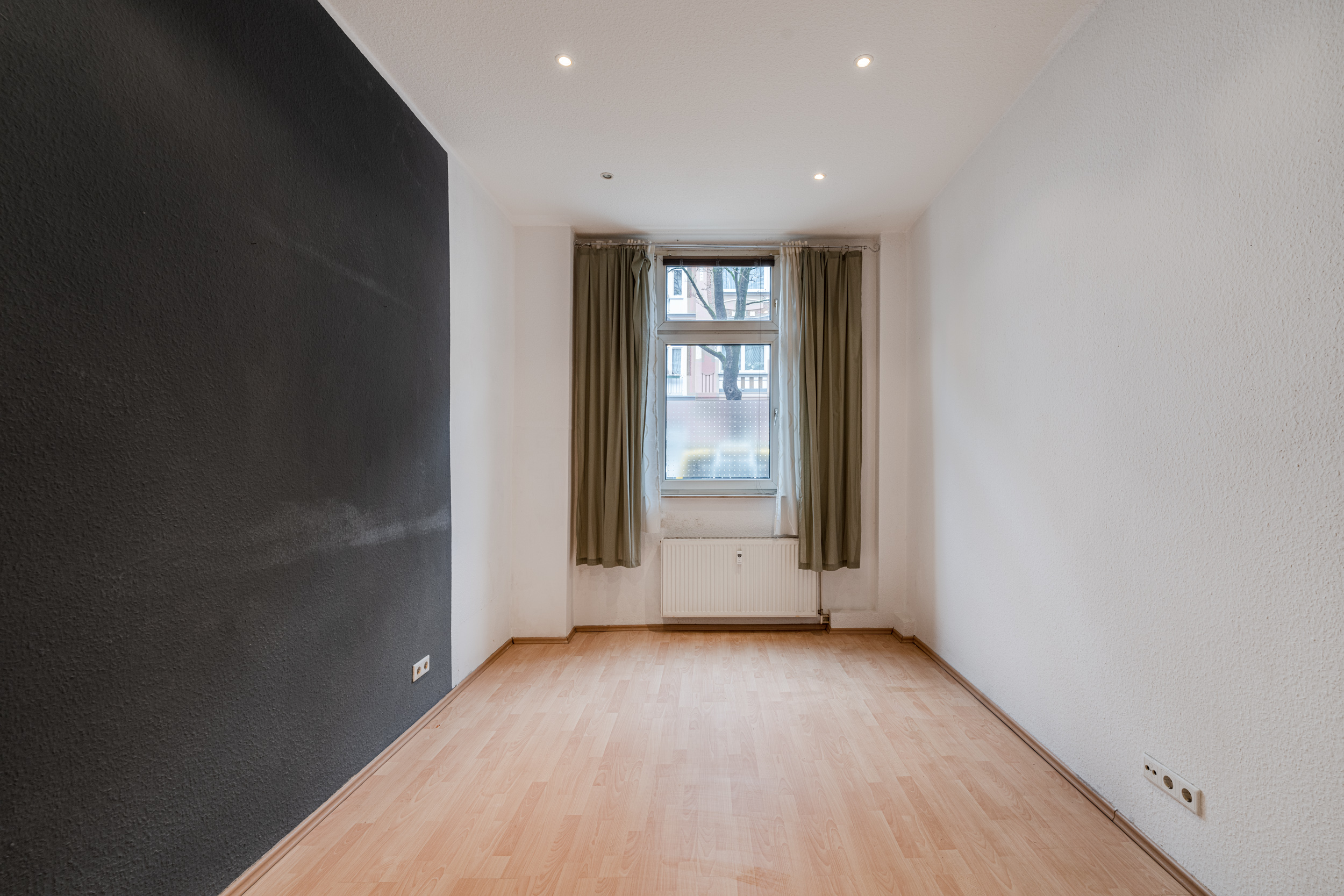 Leeres Zimmer mit hellem Laminatboden, dunkler Wand, Fenster mit Vorhängen und Heizkörper.