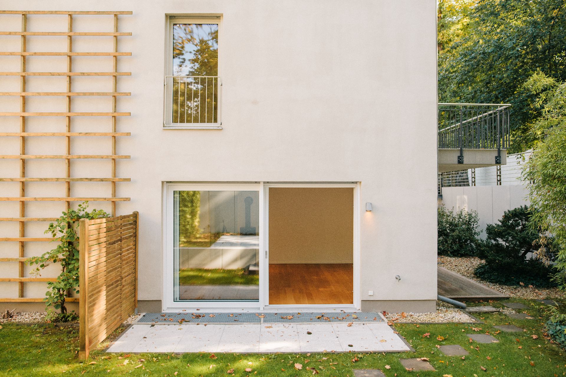 Moderne Hausfassade mit großer Glasschiebetür, kleiner Terrasse und begrüntem Garten.