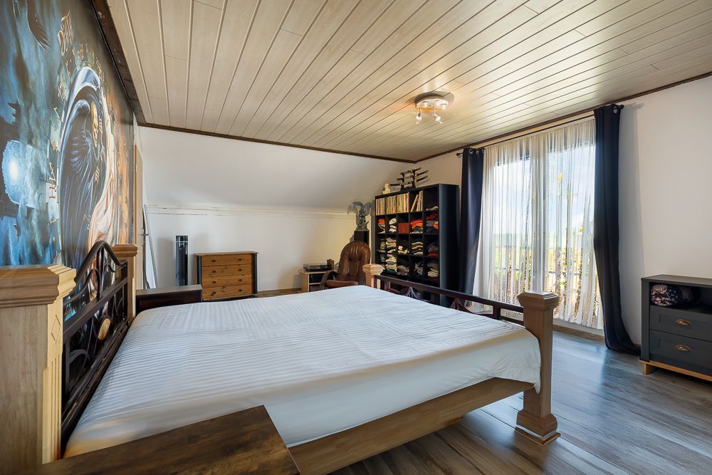 Helles Schlafzimmer mit großem Bett, Regal, Kommode und Fenster mit Vorhängen.