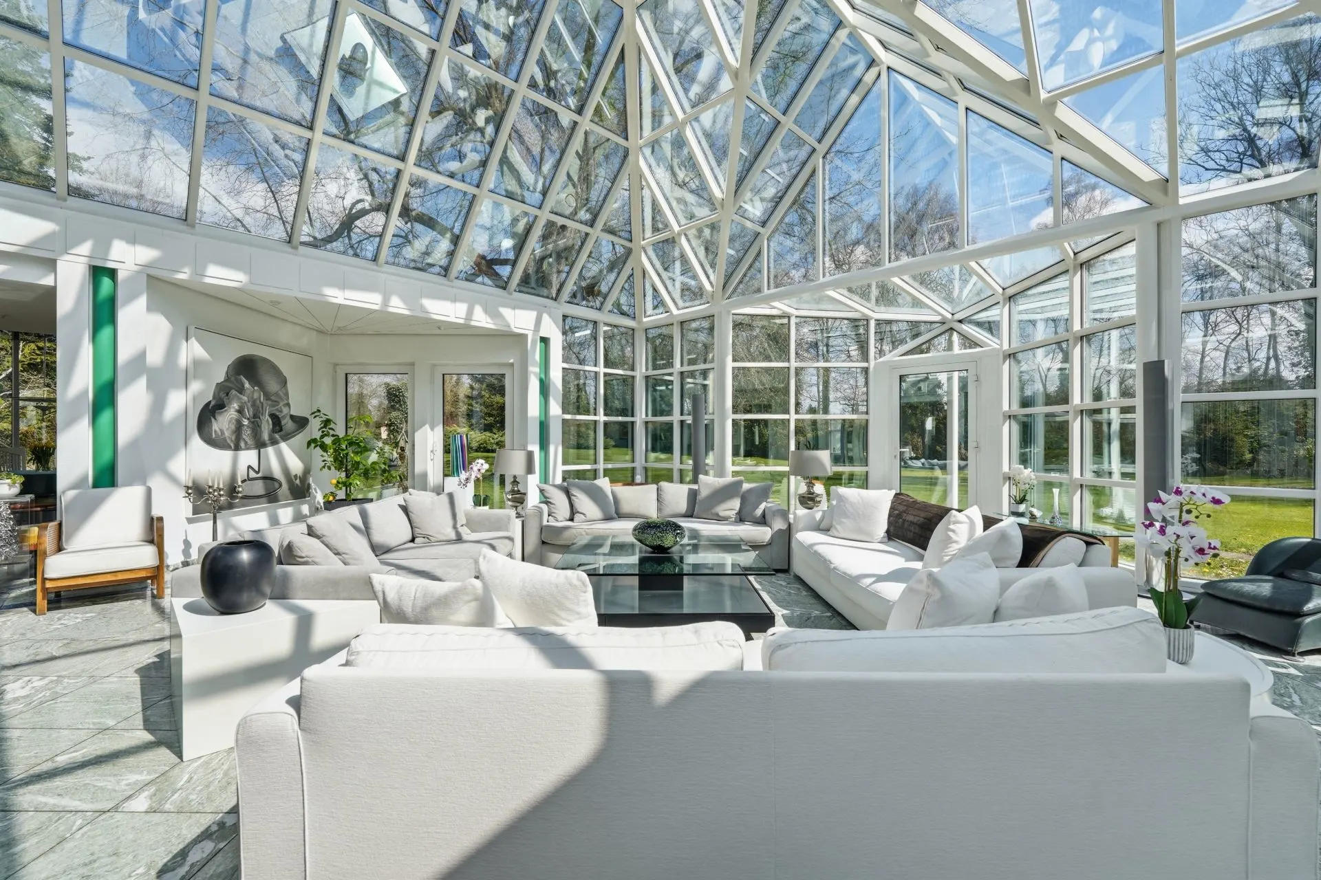 Heller Wintergarten mit Glasdach, weißen Sofas und Blick auf einen grünen Garten.