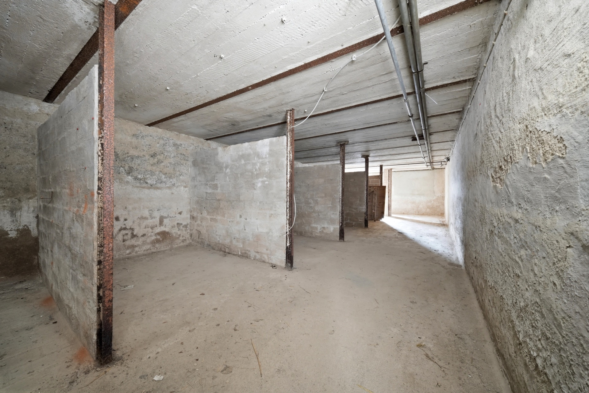 Leerer Kellerraum mit Betonwänden, Decke mit sichtbaren Rohren und mehreren Stahlstützen.
