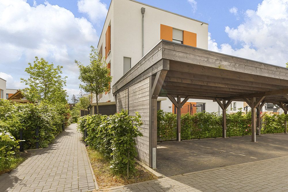 Moderne Wohngebäude mit Holz-Carport und begrünter Gehweg mit Sträuchern und Bäumen.