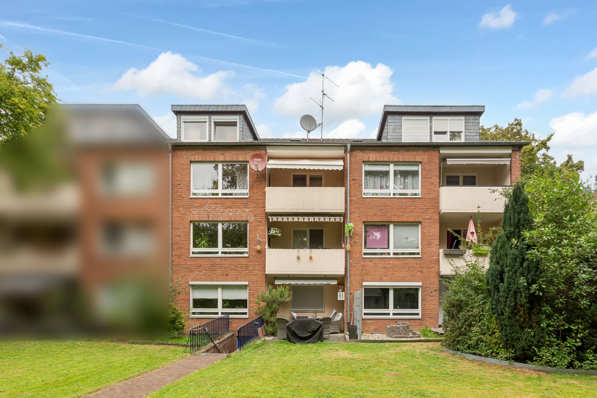 Dreistöckiges Mehrfamilienhaus mit roten Ziegeln und Balkonen, davor eine große Rasenfläche.