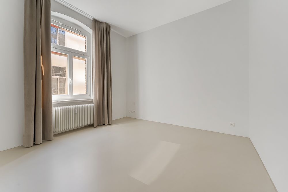 Leerer, heller Raum mit großem Fenster, beigen Vorhängen und Heizkörper darunter.