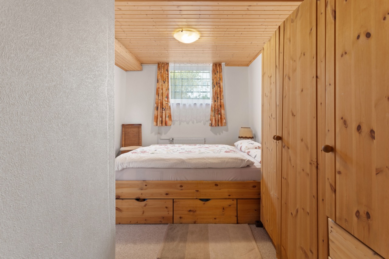 Kleines Schlafzimmer mit Holzbett, Kleiderschrank und Fenster mit Vorhängen.