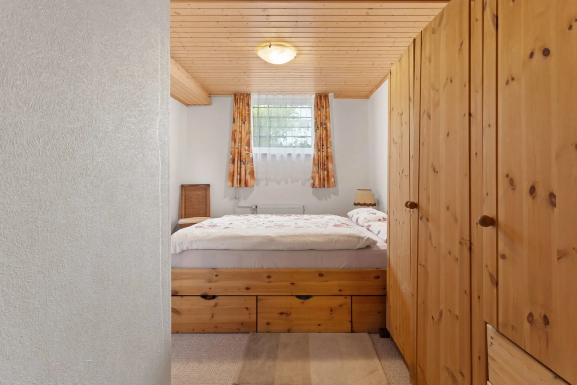 Kleines Schlafzimmer mit Holzbett, Kleiderschrank und Fenster mit Vorhängen.