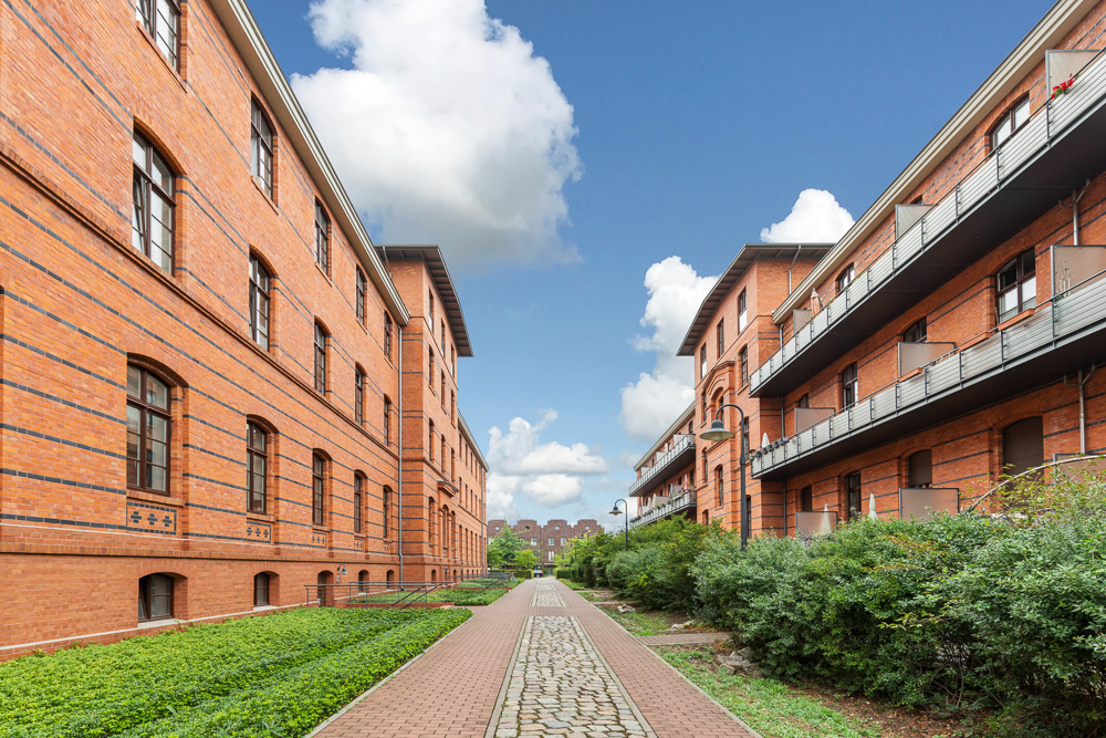 Zwei rote Backsteingebäude mit Gehweg und grünen Büschen zwischen den Häusern.