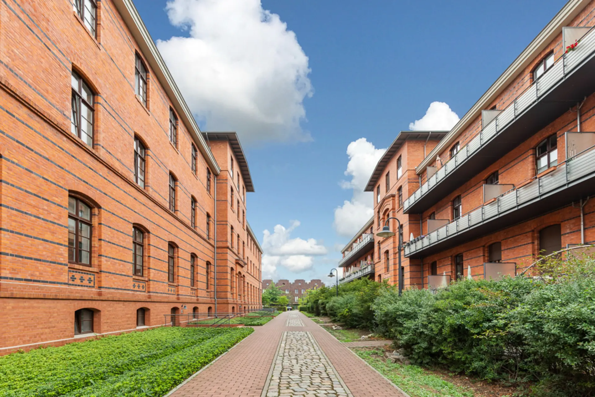 Zwei rote Backsteingebäude mit Gehweg und grünen Büschen zwischen den Häusern.